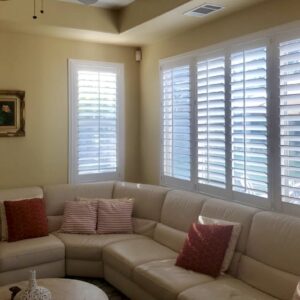 Living room comfort, beautifully illuminated! Three large windows and a corner window all feature custom white shutters over a cream sectional sofa.